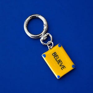 May include: A silver-toned metal keychain featuring a rectangular yellow enamel charm with blue corner accents. The word BELIEVE is printed in black capital letters on the yellow surface. The charm is attached to a circular silver split ring.