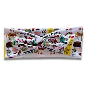 May include: A white fabric headband featuring a twisted knot design and a colourful repeating pattern of Mary Poppins themed illustrations, including umbrellas, teapots, carousel horses, and the text Mary Poppins printed in black script.