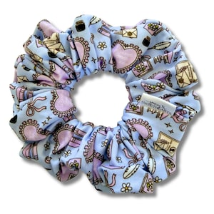 Peut inclure: Un chouchou bleu clair avec un motif répété de coeurs, d'enveloppes, de fleurs et de plumes. L'accessoire pour cheveux est en tissu doux. Le chouchou porte une étiquette indiquant "Sewing to Sweethearts".