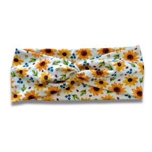 May include: A white headband with a repeating pattern of yellow sunflowers, green leaves, blue flowers, and red ladybugs. The headband is knotted at the top and is approximately 4 inches wide.