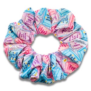 May include: A colorful scrunchie with a repeating pattern of illustrated books and text. The scrunchie features shades of pink, blue, and purple. The text includes phrases like "Things We Left Behind" and "Never Go Over".