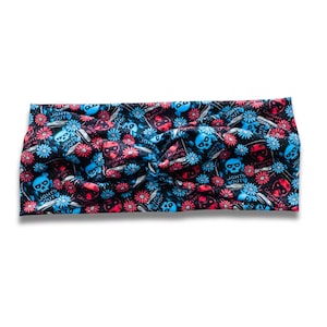 May include: A black headband featuring a repeating pattern of red and blue skulls, flowers, and the text "LIGHTS OUT" and "CAUGHT UP". The headband is twisted at the centre.