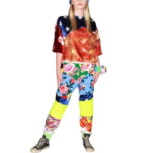 May include: A young person wearing a red, blue, and gold patterned short-sleeved shirt with a white star design. They are also wearing colorful patchwork pants with a floral print and yellow accents. The person is wearing black sneakers.