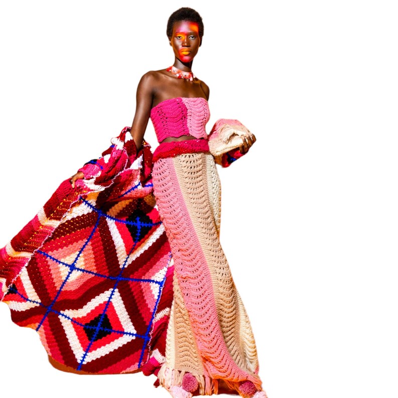 May include: A woman wearing a pink and beige crochet dress with a matching crochet shawl. The dress has a geometric pattern and the shawl has a diamond pattern. The woman is wearing a necklace and has red and orange makeup on her face.