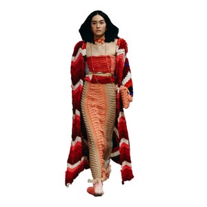 May include: A woman wearing a long, red, white, and blue crocheted cardigan over a matching crocheted skirt and top. She is wearing a red choker necklace and carrying a small crocheted bag.