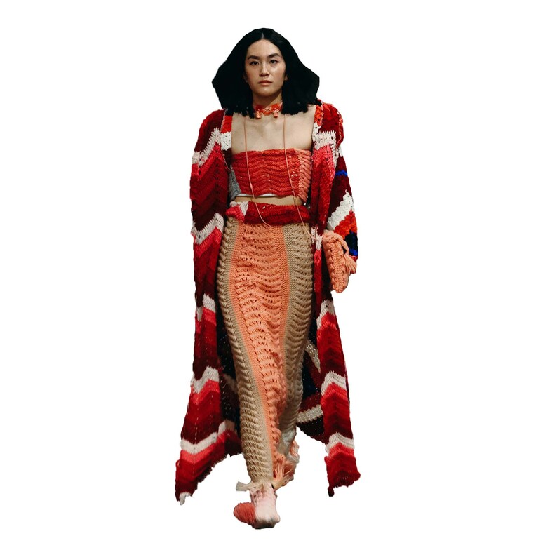 May include: A woman wearing a long, red, white, and blue crocheted cardigan over a matching crocheted skirt and top. She is wearing a red choker necklace and carrying a small crocheted bag.
