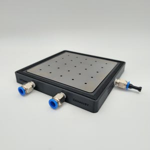 May include: A black, square vacuum chuck with a silver metal top featuring evenly spaced holes. The sides have the words "INTAKE" and "EXHAUST" printed on them. It has three silver and blue connectors.