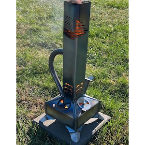 May include: A tall, square, dark metal fire pit with a cutout of an American flag on the upper side. The base features four legs and circular vents, with flames visible through the openings. A curved metal handle is attached to the side of the chimney.