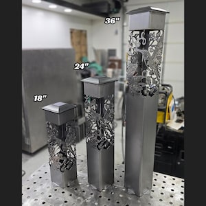 Set of three metal garden posts with hummingbird cutouts, varying in height. Displayed indoors on a workshop table with industrial equipment in the background, showcasing modern and intricate design.