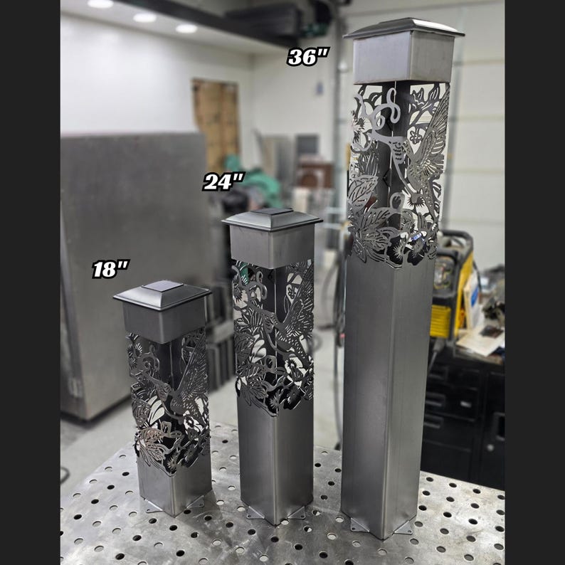 Set of three metal garden posts with hummingbird cutouts, varying in height. Displayed indoors on a workshop table with industrial equipment in the background, showcasing modern and intricate design.