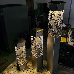 Three illuminated metal garden posts with intricate hummingbird and floral cutout designs, varying in height. Displayed on a perforated metal table in a workshop setting with dark background.