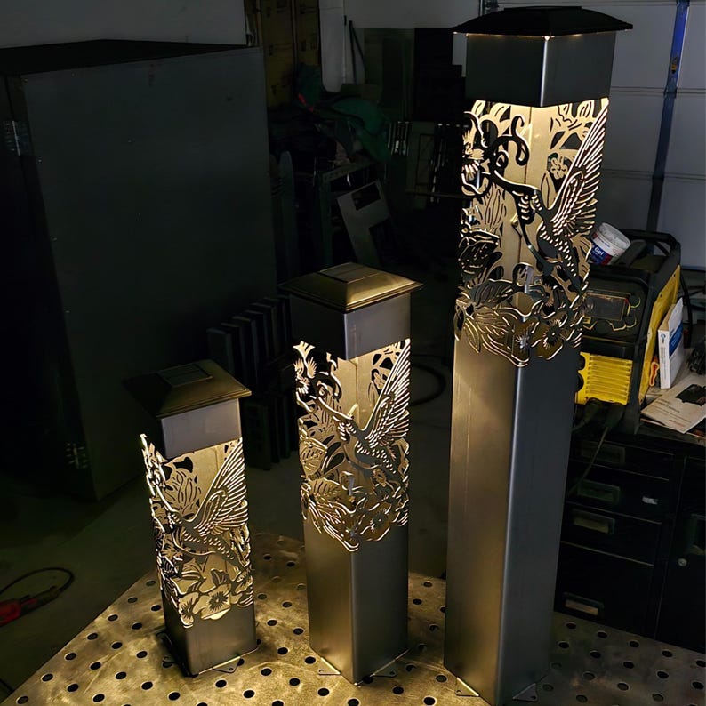 Three illuminated metal garden posts with intricate hummingbird and floral cutout designs, varying in height. Displayed on a perforated metal table in a workshop setting with dark background.