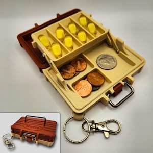 May include: A miniature tackle box-style container in tan and brown, with a hinged lid. It contains yellow capsules and coins. The container has a silver carabiner clip and a keychain. The word "TACKLE" is printed on the side.