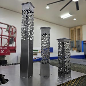 Puede incluir: Tres pilares decorativos de metal de diferentes alturas. Cada pilar es cuadrado con una parte superior plana y está adornado con recortes en forma de corazón. El metal tiene un acabado plateado cepillado. Los pilares se apoyan sobre una superficie metálica.