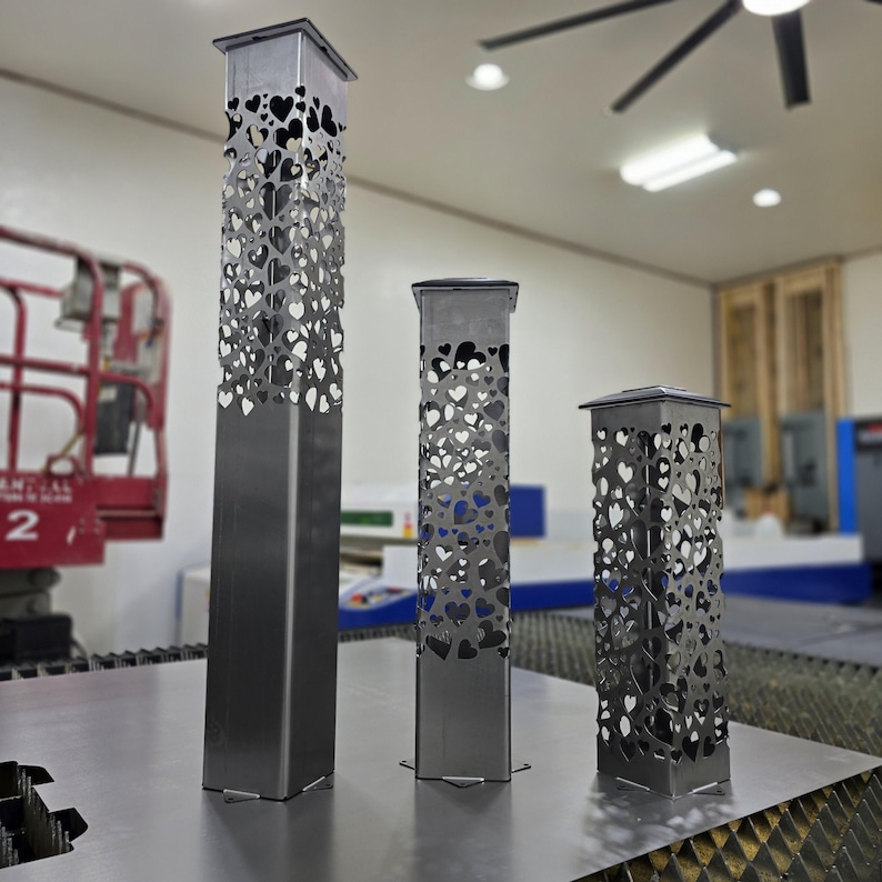 Set of three metal garden posts with heart-shaped cutouts, varying in height. Displayed indoors on a workshop table with industrial equipment in the background, showcasing modern and intricate design.