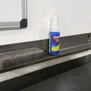 May include: A metal shelf with a black finish is mounted to a white wall. A blue spray bottle labeled "Exacto" is on the shelf, along with a gray rectangular eraser.