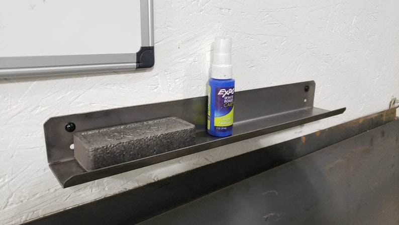 May include: A metal shelf with a black finish is mounted to a white wall. A blue spray bottle labeled "Exacto" is on the shelf, along with a gray rectangular eraser.