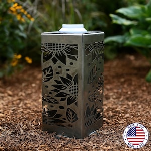 May include: A rectangular metal garden lantern with a white top and sunflower cutouts. The lantern is made in the USA and sits on a bed of brown mulch. The sunflower design is visible on all four sides of the lantern.