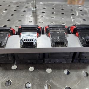 May include: A metal rack with four battery packs. The battery packs are black with red accents and have a silver connector. The rack is made of metal and has a silver finish.