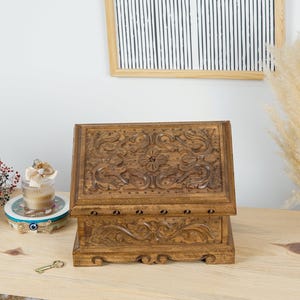 Peut inclure: Une boîte à bijoux en bois avec des sculptures florales complexes. La boîte est rectangulaire avec un couvercle à charnière et un trou de serrure. La boîte est de couleur marron chaud et repose sur une étagère en bois.