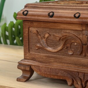 Op de afbeelding: Een houten doos met ingewikkelde houtsnijwerk en decoratieve metalen accenten. De doos heeft een donkerbruine afwerking en staat op een houten oppervlak.