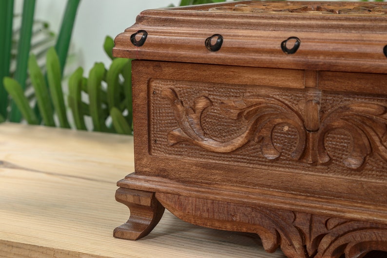 Op de afbeelding: Een houten doos met ingewikkelde houtsnijwerk en decoratieve metalen accenten. De doos heeft een donkerbruine afwerking en staat op een houten oppervlak.