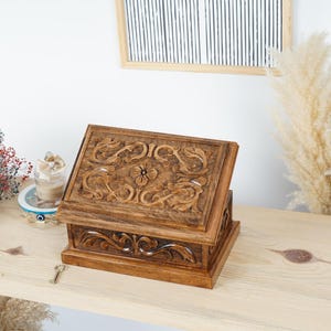 May include: A wooden jewellery box with detailed floral carvings on the lid and sides. The box is a warm brown colour and has a hinged lid. A small key rests near the box. The box is on a wooden surface.