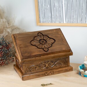 May include: A rectangular wooden box with a dark brown finish, featuring a carved floral motif on the slanted lid and intricate scrollwork carvings along the front panel. The box has a stepped base design and is crafted from natural wood grain.