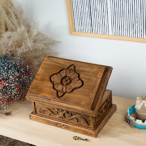 May include: A rectangular wooden box with a hinged lid featuring a carved floral motif on top and intricate scrollwork patterns along the sides. The box is finished in a medium brown wood stain and includes a small metal key placed in front.