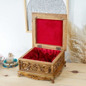 Peut inclure: Une boîte carrée en bois avec un couvercle articulé ouvert, présentant des motifs floraux sculptés complexes sur les côtés extérieurs et quatre petits pieds. L'intérieur de la boîte et le dessous du couvercle sont doublés de velours rouge.