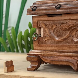 Op de afbeelding: Een houten kist met ingewikkelde houtsnijwerk en een sleutelgat. De kist is gesloten en heeft een donkerbruine afwerking. De kist staat op een houten oppervlak.