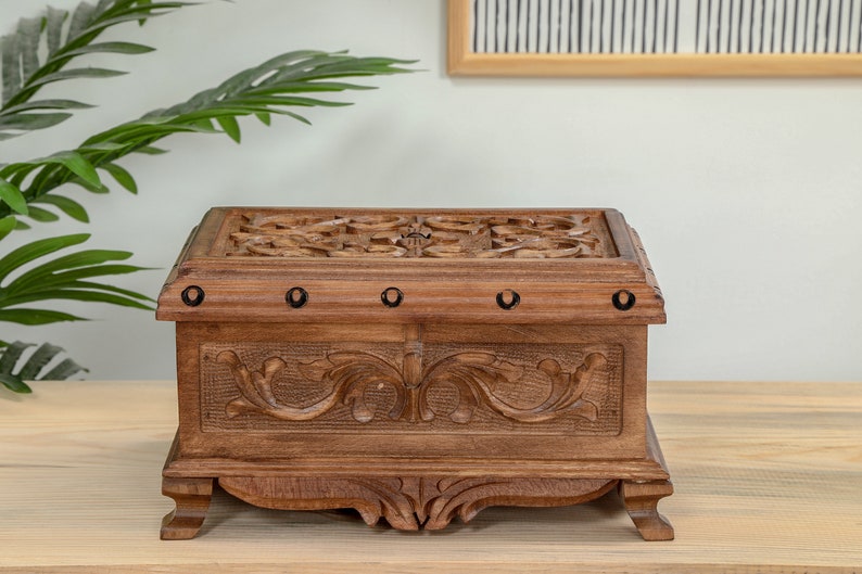 Op de afbeelding: Een houten kist met ingewikkelde houtsnijwerk en metalen accenten. De kist heeft een scharnierend deksel en staat op vier poten.