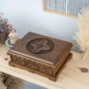May include: A dark brown wooden box with carved floral details and a starburst design on the lid. The rectangular box is displayed on a light wooden surface. The background features decorative elements. The box is approximately 25 cm long.