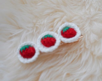 Puede incluir: Tres pinzas para el pelo de fresa tejidas a crochet con hilo blanco y detalles rojos y verdes. Las pinzas están unidas a una barra de metal dorada.