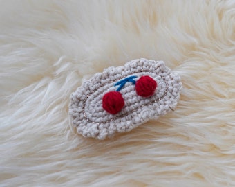 Puede incluir: Una pinza para el pelo ovalada de crochet blanco con dos cerezas rojas bordadas.