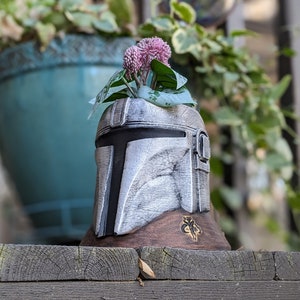 May include: A small potted plant with pink flowers growing out of a ceramic Mandalorian helmet planter. The planter is painted in a silver and black color scheme and has a brown base.