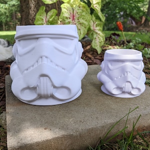 May include: Two white ceramic planters shaped like the head of a Stormtrooper from Star Wars. The larger planter is in the foreground and the smaller planter is in the background.