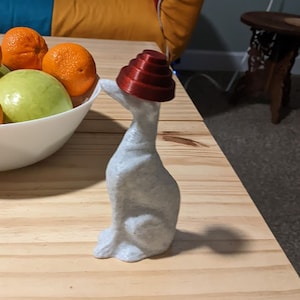 Puede incluir: Una figurita de cerámica blanca de un perro con un sombrero rojo y blanco. El perro está sentado en una mesa de madera con un tazón de naranjas y una manzana verde.