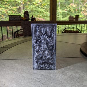 May include: A gray and white 3D printed model of a character frozen in carbonite. The model is a replica of the iconic scene from the Star Wars movie.