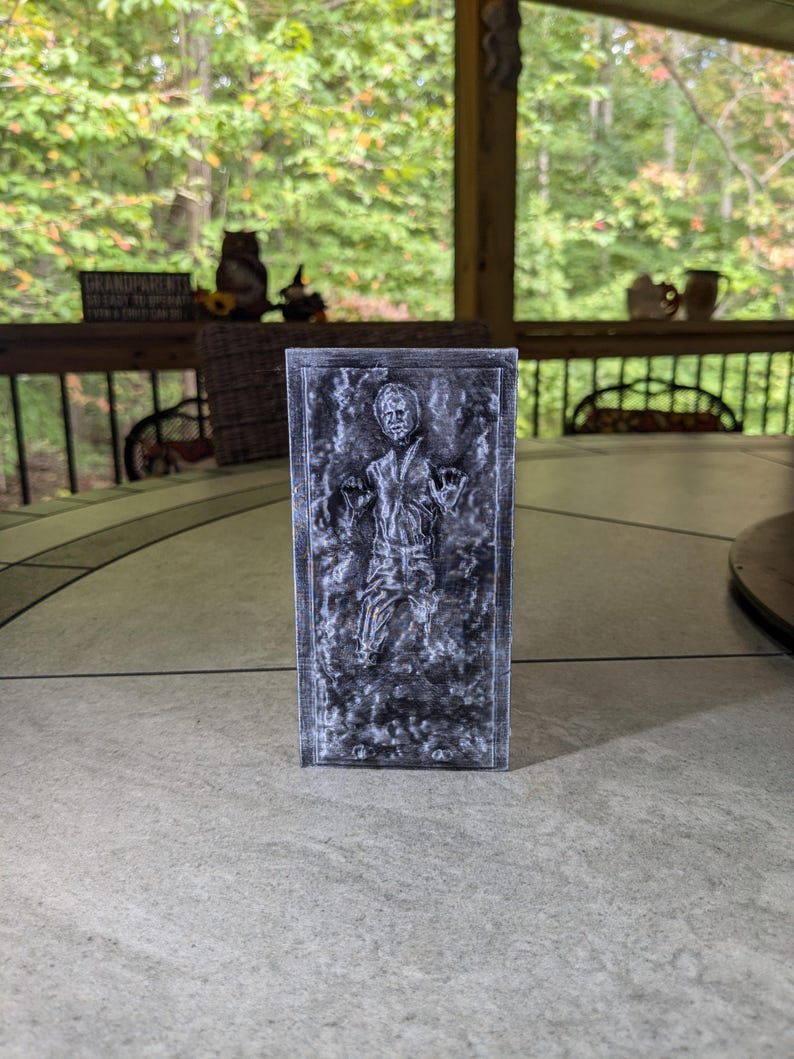 May include: A gray and white 3D printed model of a character frozen in carbonite. The model is a replica of the iconic scene from the Star Wars movie.