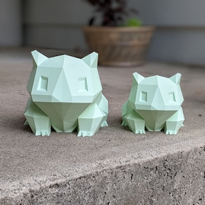 Può includere: Due figurine geometriche di colore verde, stampate in 3D, di un personaggio Pokemon. La figurina più grande è in primo piano e quella più piccola è sullo sfondo.
