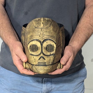 May include: A gold and black ceramic planter shaped like a robot head. The planter has a detailed design with geometric patterns and a textured surface.