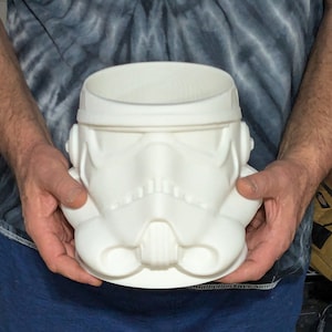 Può includere: Un contenitore bianco a forma di casco da Stormtrooper. Il contenitore è una replica dettagliata del casco dell'iconico personaggio di Star Wars, con i tratti distintivi del viso e la forma del casco. Il contenitore è tenuto da una persona.