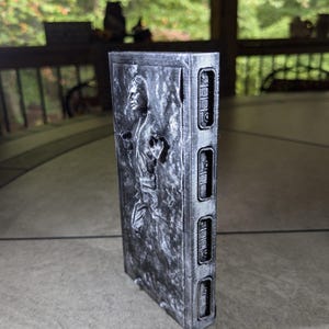 May include: A silver-colored 3D printed model of Han Solo frozen in carbonite. The model is detailed with a textured surface and a raised image of Han Solo's face. The model is approximately 15 cm tall.