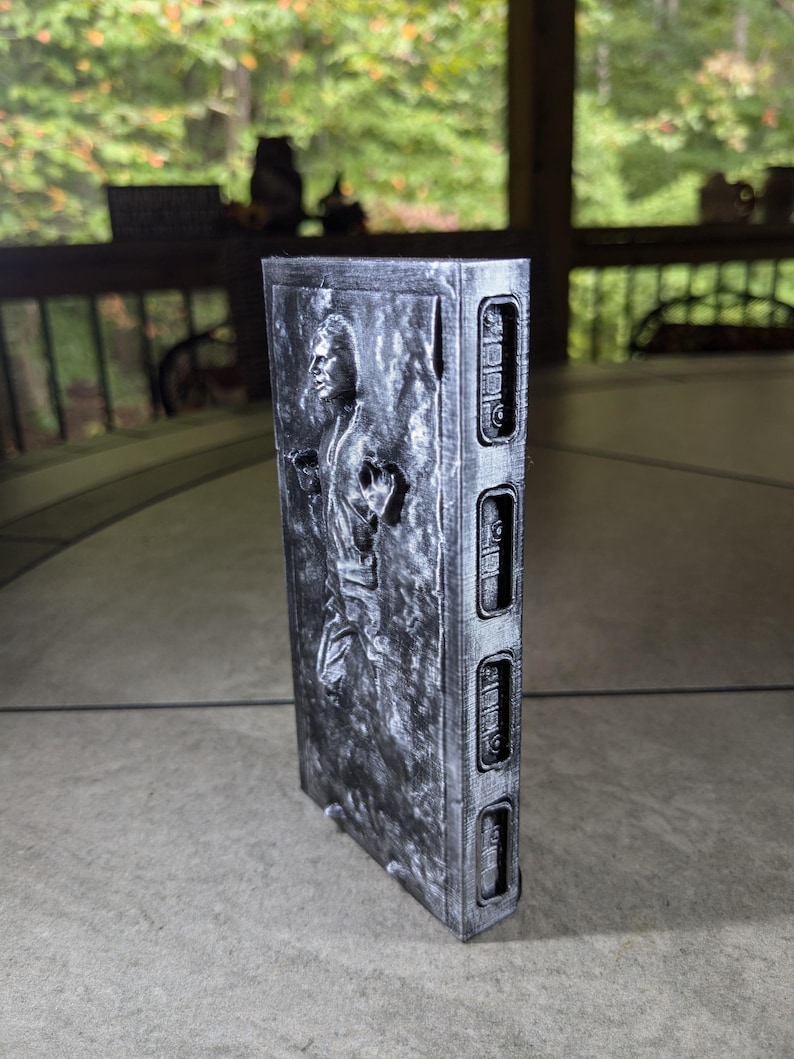 May include: A silver-colored 3D printed model of Han Solo frozen in carbonite. The model is detailed with a textured surface and a raised image of Han Solo's face. The model is approximately 15 cm tall.