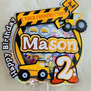Könnte beinhalten: Ein gelber und schwarzer Kuchentopper zum Thema Bau mit dem Namen "Mason" und der Zahl "2" darauf. Der Topper zeigt einen gelben Kipplaster, einen Baukran und ein Schild mit der Aufschrift "Im Bau".