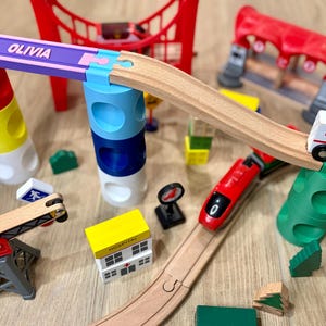 May include: A wooden toy train set with a red train, an ambulance, and a bridge. The set includes a track, a hospital, and a tower with the name "Olivia" on it. The set is made of wood and plastic.