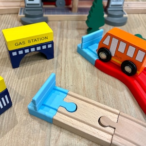 May include: Wooden toy train set with a gas station, bus, and track pieces. The gas station is yellow and blue, the bus is orange, and the tracks are light brown. The set includes a green tree and a train station.