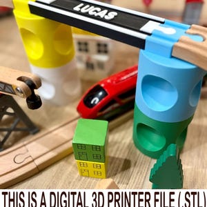 May include: A 3D printed toy train bridge support system featuring a black track segment with the name LUCAS in white letters, supported by yellow and blue cylindrical pillars, alongside a red train and small wooden building blocks.