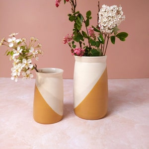 Peut inclure: Deux vases en céramique avec un glaçage blanc et jaune. Le vase le plus grand est rempli de fleurs blanches, et le vase le plus petit est rempli de fleurs roses.